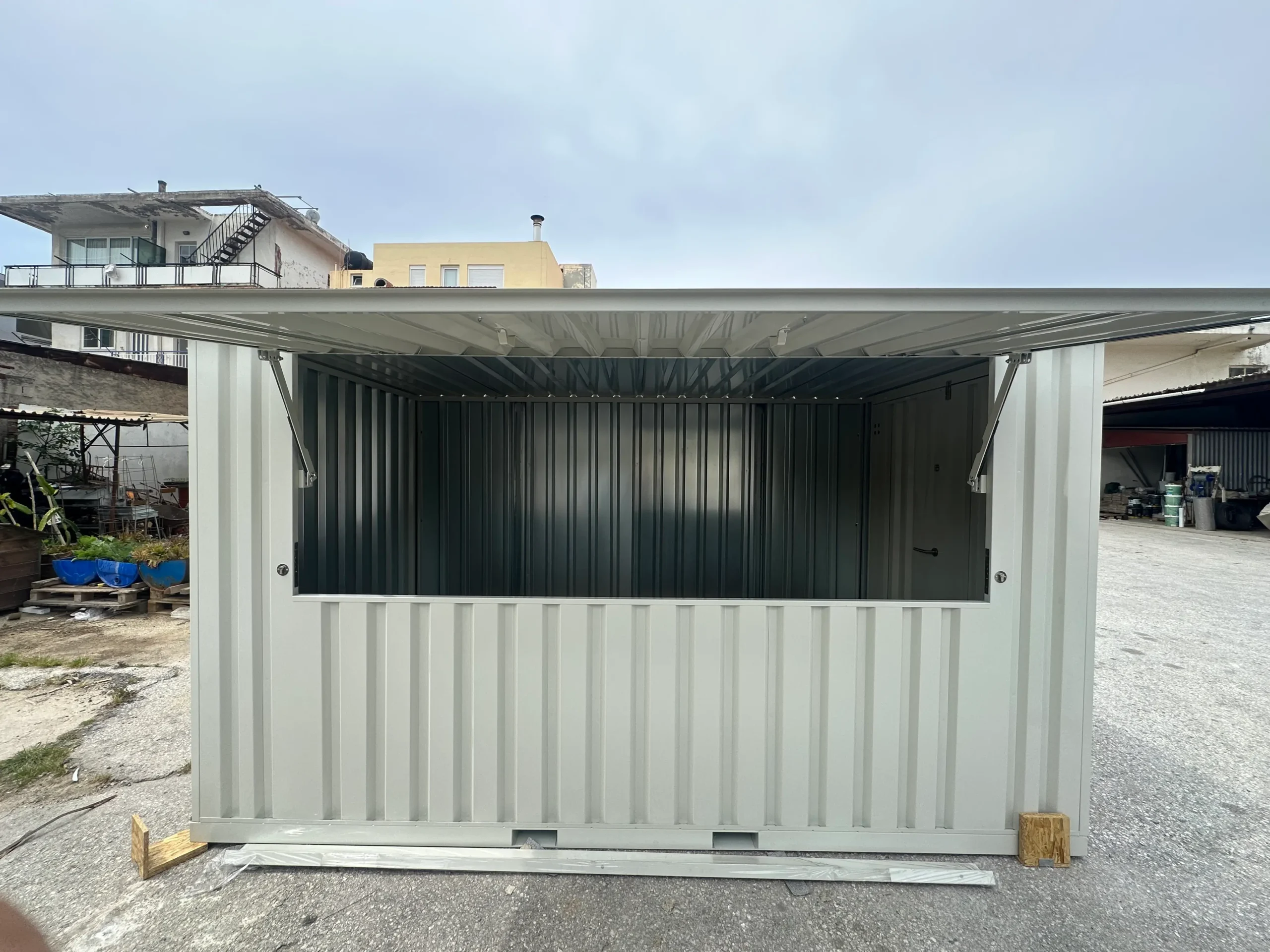 A grey shipping container modified with a large lift-up serving window, placed in an outdoor industrial area.