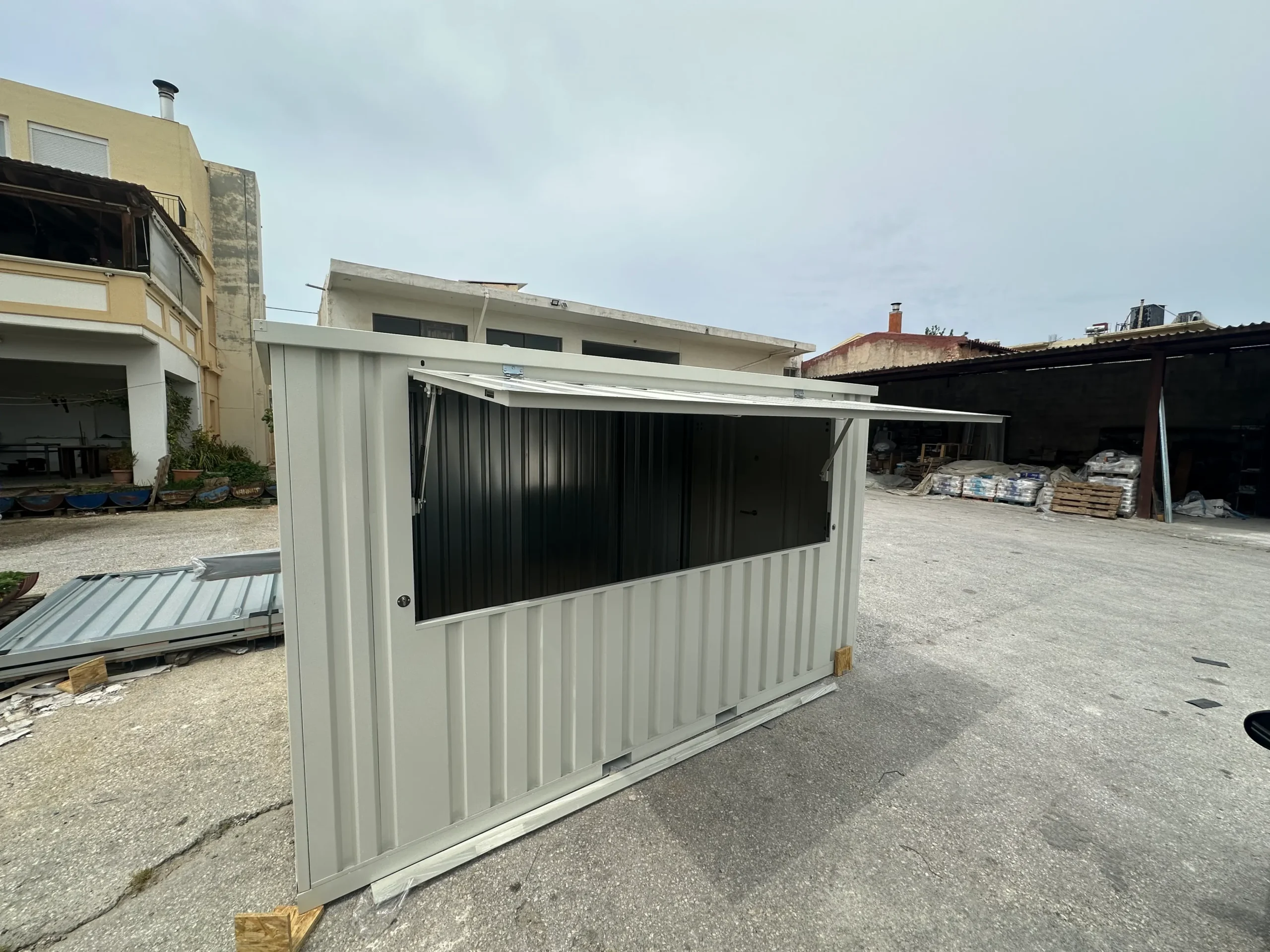A grey shipping container modified with a large lift-up serving window, placed in an outdoor industrial area.