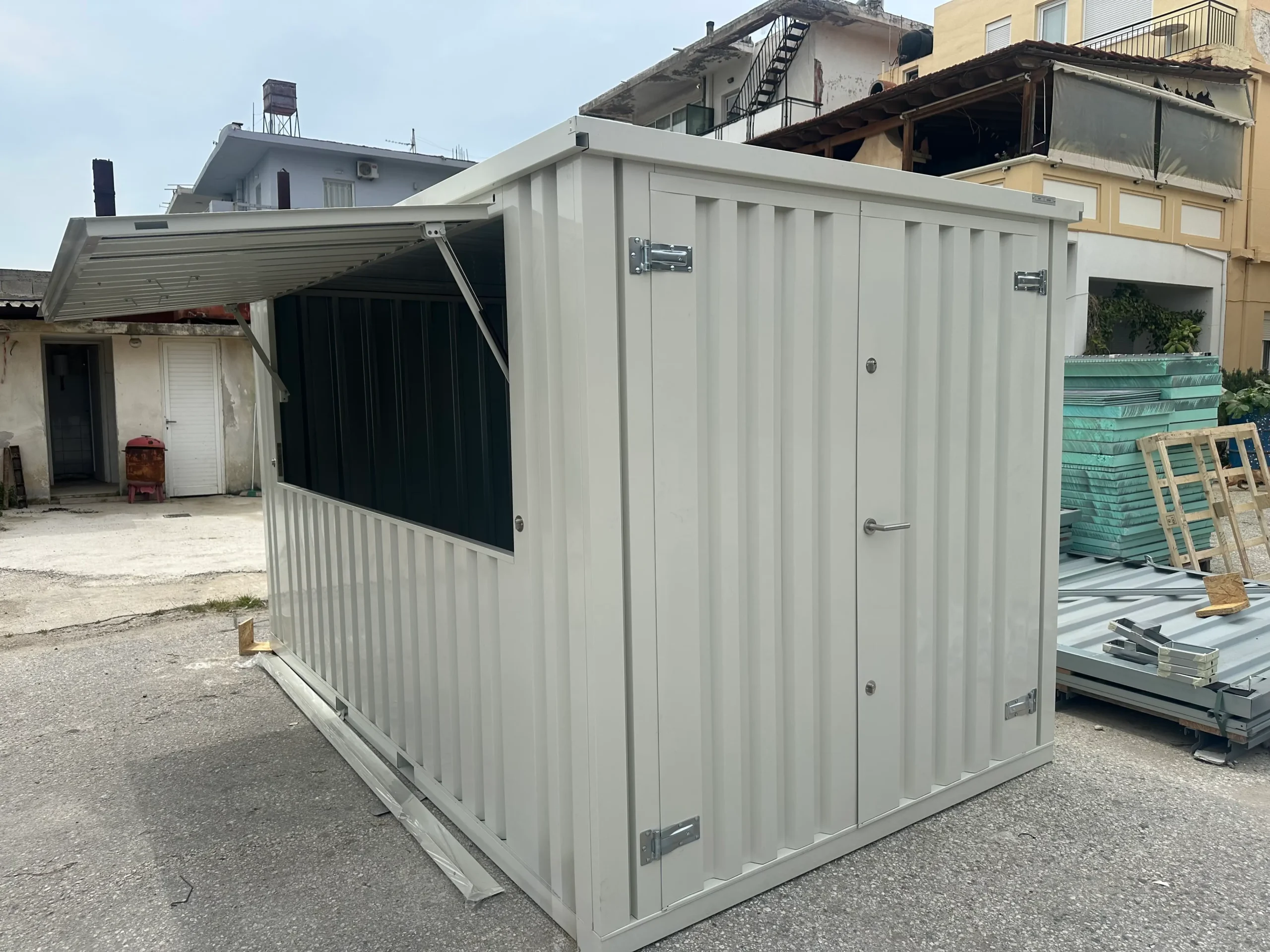 A grey shipping container modified with a large lift-up serving window, placed in an outdoor industrial area.